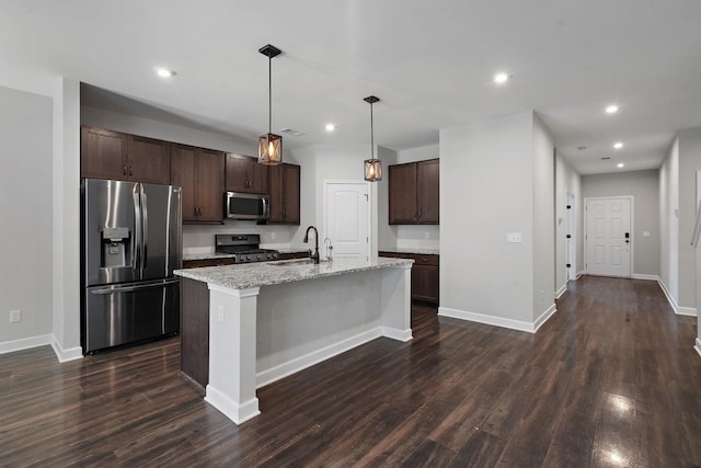 kitchen featuring appliances with stainless steel finishes, hanging light fixtures, dark brown cabinets, an island with sink, and light stone counters