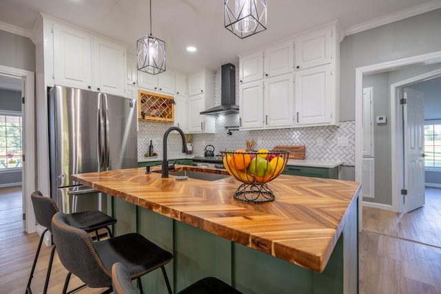 kitchen featuring wall chimney range hood, wooden counters, stainless steel refrigerator, white cabinets, and a center island with sink