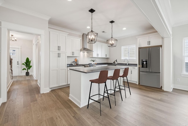 kitchen with stainless steel refrigerator with ice dispenser, pendant lighting, wall chimney exhaust hood, sink, and white cabinetry