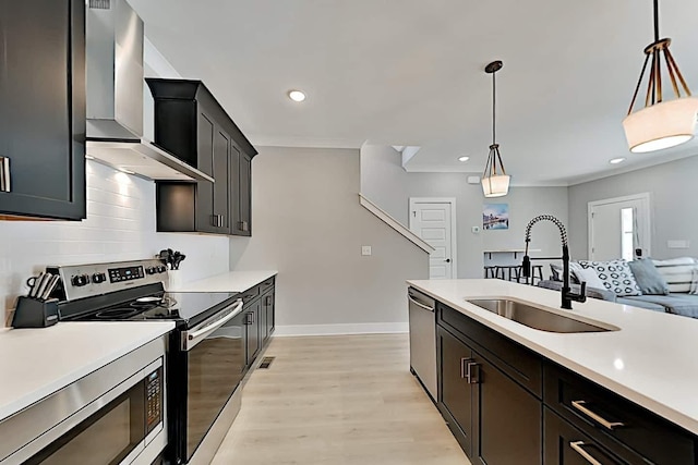 kitchen with wall chimney range hood, stainless steel appliances, sink, hanging light fixtures, and light hardwood / wood-style flooring