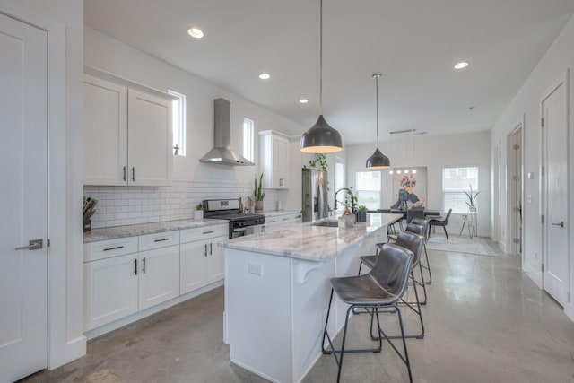 kitchen with wall chimney exhaust hood, white cabinets, gas stove, and an island with sink