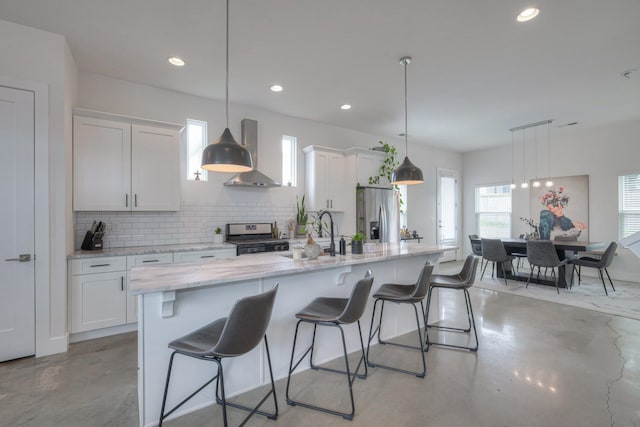 kitchen with a large island with sink, appliances with stainless steel finishes, pendant lighting, wall chimney exhaust hood, and white cabinetry