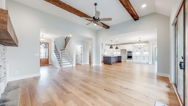 unfurnished living room featuring light hardwood / wood-style flooring, sink, a stone fireplace, ceiling fan with notable chandelier, and beamed ceiling