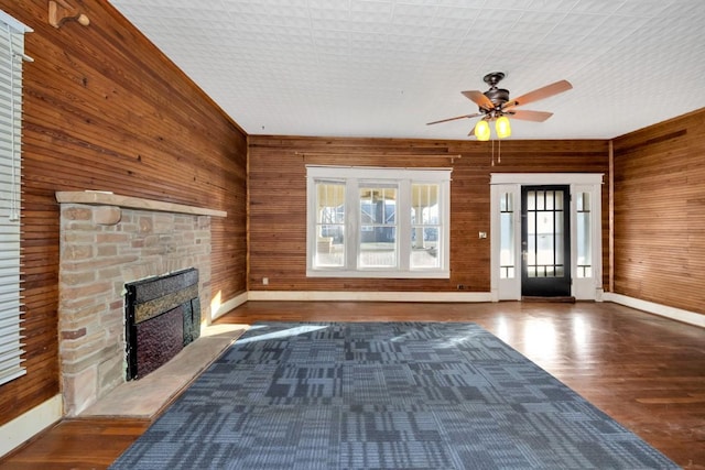 unfurnished living room with ceiling fan, a stone fireplace, wood walls, and dark hardwood / wood-style flooring