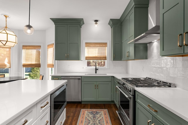 kitchen featuring pendant lighting, appliances with stainless steel finishes, wall chimney exhaust hood, sink, and green cabinetry