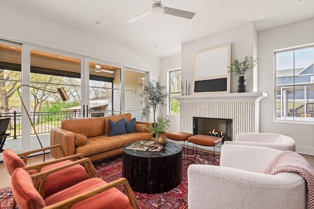 living area featuring baseboards, a warm lit fireplace, wood finished floors, and a ceiling fan