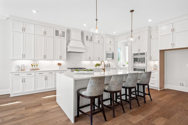 kitchen with premium range hood, appliances with stainless steel finishes, pendant lighting, white cabinets, and a center island with sink