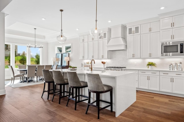 kitchen with a breakfast bar, premium range hood, a kitchen island with sink, stainless steel microwave, and white cabinets