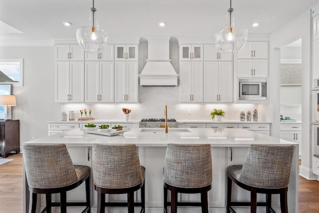 kitchen with white cabinets, stainless steel microwave, custom range hood, and a center island with sink