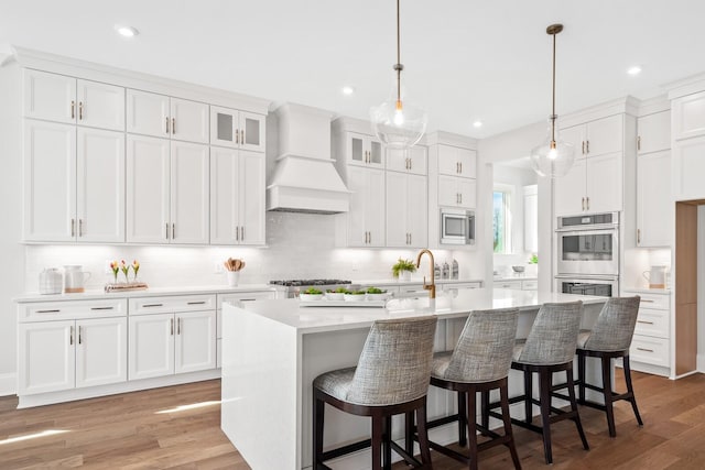 kitchen with pendant lighting, an island with sink, white cabinets, custom exhaust hood, and stainless steel appliances