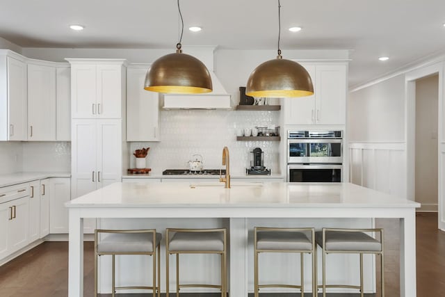 kitchen featuring white cabinets, hanging light fixtures, a kitchen island with sink, and stainless steel double oven