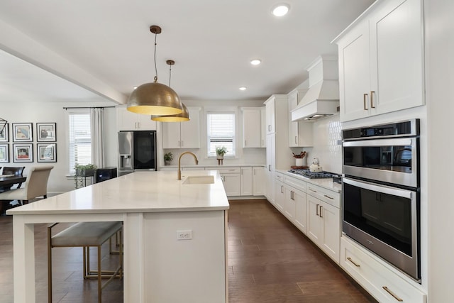 kitchen with custom exhaust hood, decorative light fixtures, stainless steel appliances, sink, and a kitchen island with sink