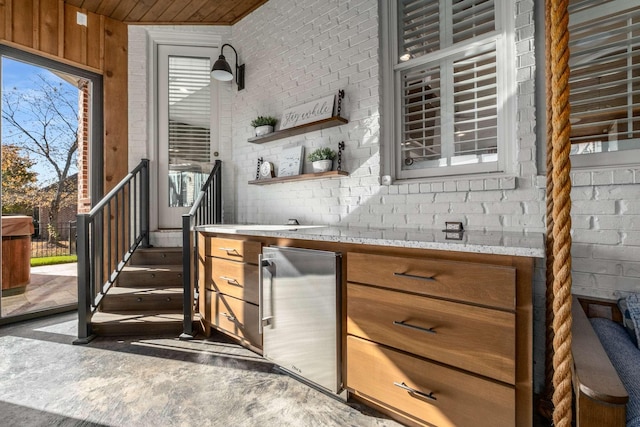 kitchen with brown cabinets, stainless steel refrigerator, light stone counters, open shelves, and brick wall