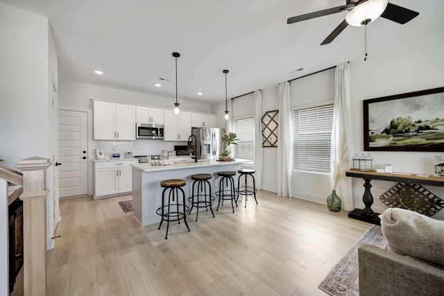 kitchen with hanging light fixtures, stainless steel appliances, tasteful backsplash, white cabinets, and a center island with sink