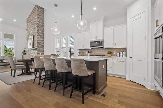 kitchen featuring an island with sink, white cabinets, decorative backsplash, hanging light fixtures, and stainless steel appliances