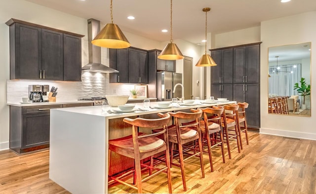 kitchen with stainless steel refrigerator with ice dispenser, wall chimney exhaust hood, a breakfast bar, decorative light fixtures, and a kitchen island with sink