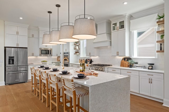 kitchen with white cabinetry, custom exhaust hood, a kitchen island with sink, stainless steel appliances, and light hardwood / wood-style flooring