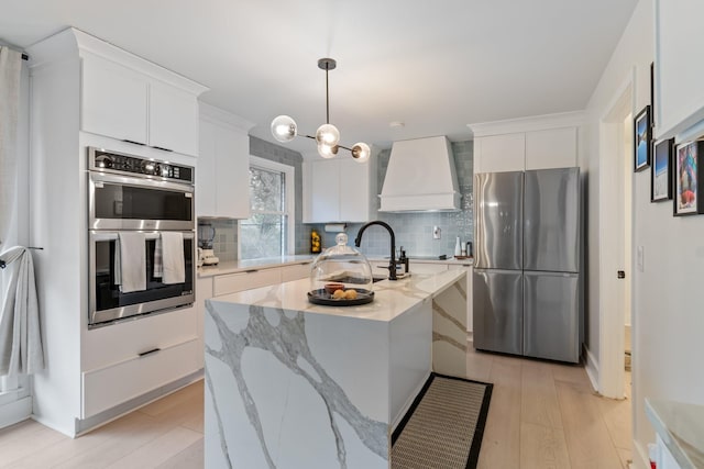 kitchen featuring pendant lighting, appliances with stainless steel finishes, white cabinets, a center island with sink, and custom exhaust hood