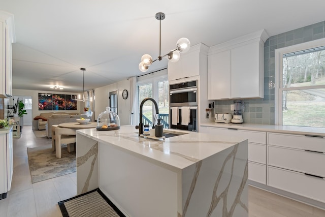 kitchen featuring double oven, white cabinetry, sink, light stone counters, and a center island with sink