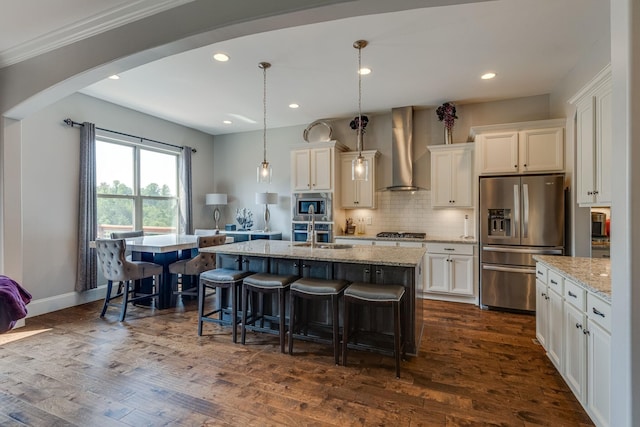 kitchen featuring hanging light fixtures, stainless steel appliances, an island with sink, white cabinets, and wall chimney exhaust hood