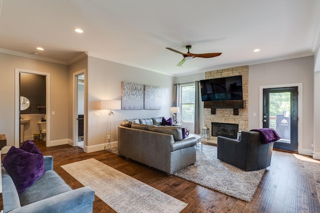 living room featuring dark wood-type flooring, ceiling fan, crown molding, and a stone fireplace