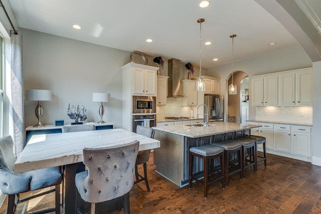 kitchen with appliances with stainless steel finishes, a kitchen bar, hanging light fixtures, a center island with sink, and wall chimney exhaust hood