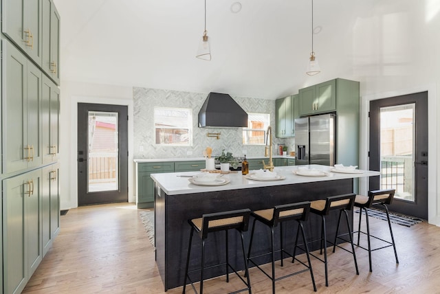 kitchen with pendant lighting, wall chimney exhaust hood, stainless steel fridge, and a kitchen island with sink