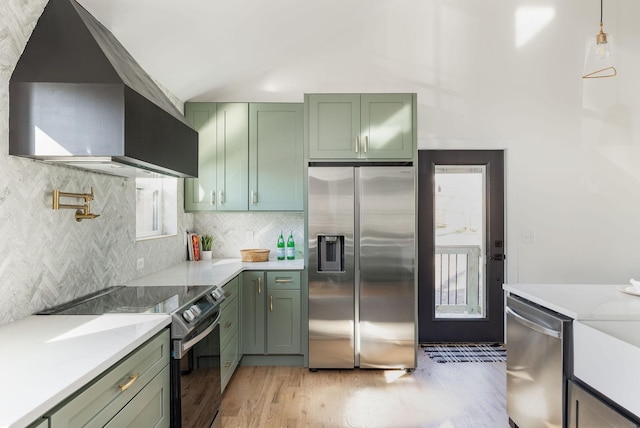 kitchen featuring wall chimney range hood, green cabinets, appliances with stainless steel finishes, hanging light fixtures, and light stone counters