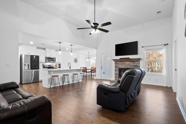 living room featuring dark wood-type flooring, ceiling fan, a stone fireplace, and a high ceiling