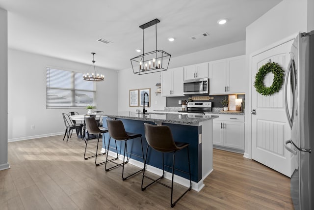 kitchen featuring appliances with stainless steel finishes, decorative light fixtures, white cabinetry, an island with sink, and dark stone counters