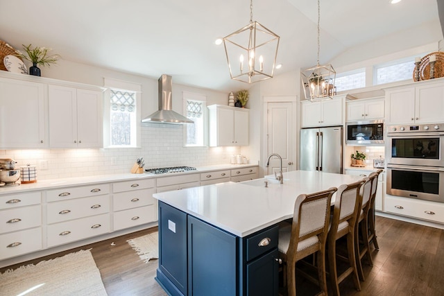 kitchen with sink, wall chimney range hood, stainless steel appliances, a kitchen island with sink, and white cabinets