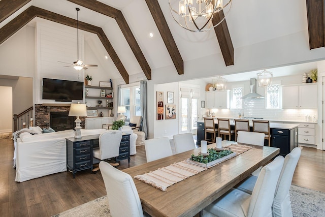 dining area featuring dark wood-type flooring, ceiling fan with notable chandelier, high vaulted ceiling, a stone fireplace, and beamed ceiling