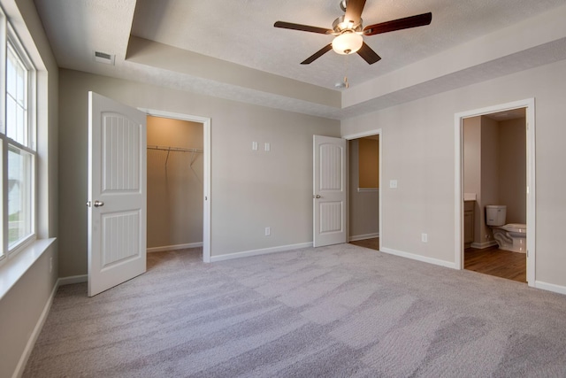 unfurnished bedroom with a tray ceiling, ensuite bath, light colored carpet, and ceiling fan