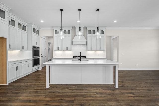 kitchen with pendant lighting, custom range hood, white cabinetry, and an island with sink