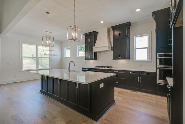 kitchen with sink, appliances with stainless steel finishes, a kitchen island with sink, hanging light fixtures, and custom exhaust hood