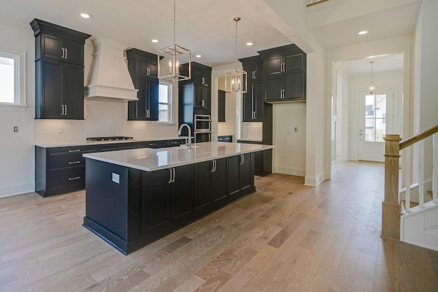 kitchen with appliances with stainless steel finishes, decorative light fixtures, custom range hood, a center island with sink, and light wood-type flooring