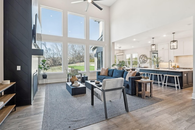 living room with sink, light wood-type flooring, a fireplace, ceiling fan with notable chandelier, and a high ceiling