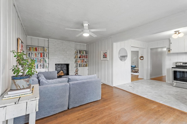 living room with built in shelves, a stone fireplace, light hardwood / wood-style flooring, a textured ceiling, and ceiling fan