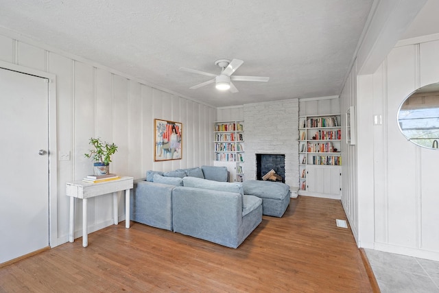 living room featuring built in features, ceiling fan, a textured ceiling, a stone fireplace, and light wood-type flooring