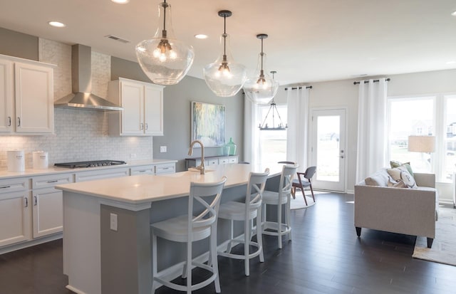 kitchen featuring stainless steel gas stovetop, white cabinetry, backsplash, a kitchen island with sink, and wall chimney range hood