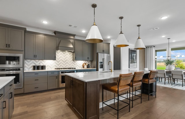 kitchen featuring premium range hood, tasteful backsplash, decorative light fixtures, appliances with stainless steel finishes, and a kitchen island with sink