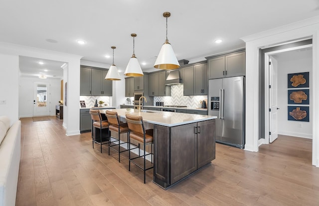 kitchen featuring a breakfast bar, an island with sink, high end refrigerator, hanging light fixtures, and wall chimney range hood