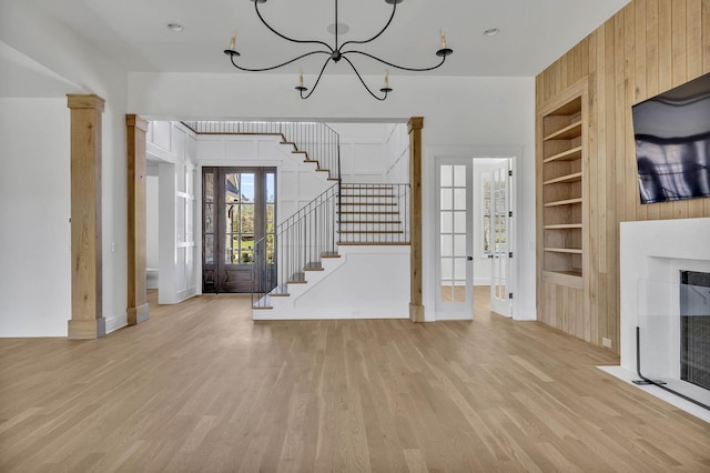 unfurnished living room with built in features, wooden walls, light hardwood / wood-style floors, french doors, and a chandelier