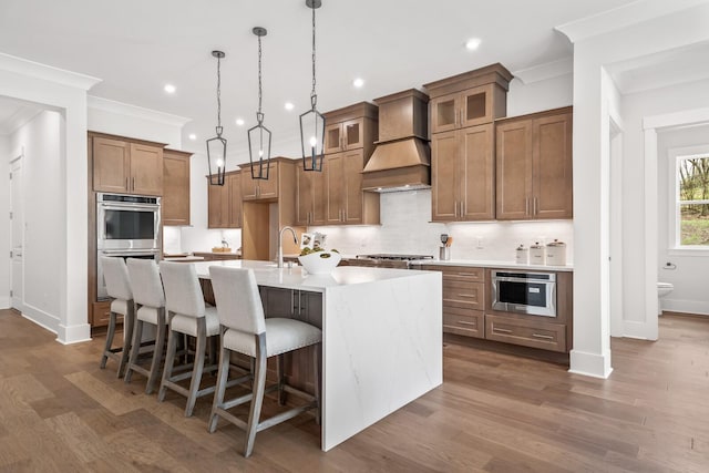 kitchen with appliances with stainless steel finishes, dark hardwood / wood-style floors, decorative light fixtures, an island with sink, and custom range hood