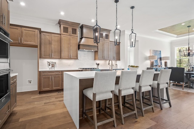 kitchen with hardwood / wood-style floors, decorative light fixtures, a kitchen island with sink, crown molding, and custom range hood