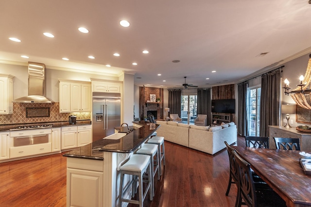 kitchen featuring built in fridge, crown molding, wall chimney range hood, and a fireplace