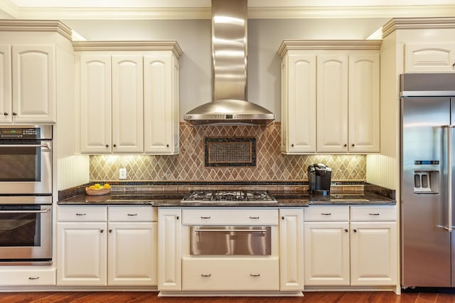 kitchen featuring wall chimney exhaust hood, crown molding, tasteful backsplash, appliances with stainless steel finishes, and dark stone counters