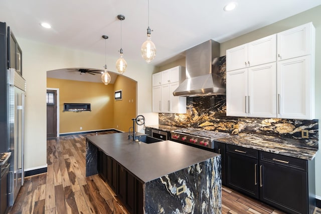 kitchen featuring white cabinetry, an island with sink, sink, hanging light fixtures, and wall chimney exhaust hood