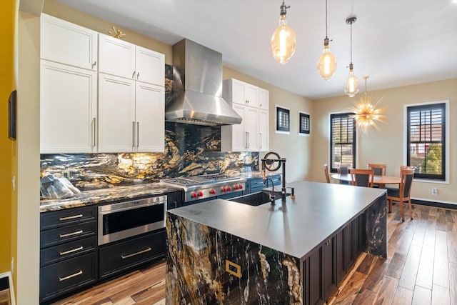 kitchen with sink, hanging light fixtures, a center island with sink, wall chimney range hood, and white cabinets