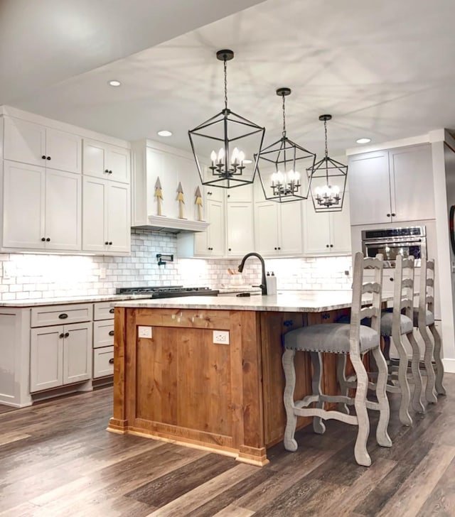 kitchen featuring white cabinetry, light stone counters, an island with sink, decorative light fixtures, and oven